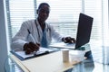 Male doctor writing on a clipboard while using laptop at desk Royalty Free Stock Photo