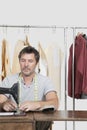 Male dressmaker stitching cloth on sewing machine with clothes rack in background Royalty Free Stock Photo