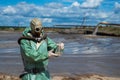 A male environmentalist in a green protective suit and gas mask takes a sample of water in a polluted lake for research