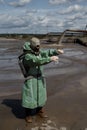 A male environmentalist in a green protective suit and gas mask takes a sample of water in a polluted lake. Waste from