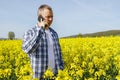 A male farmer agronomist stands in a field of flowering rapeseed and talks on the phone. The farmer checks the fields