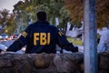 Male FBI agent wearing dark blue coat with FBI logo looking down the street with cars in the dusk seen from behind