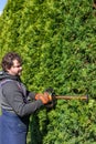 Male gardener in uniform using electric hedge cutter for work outdoors. Caucasian man shaping overgrown thuja during summer time Royalty Free Stock Photo