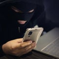 Male hacker in a black mask uses smartphone and laptop. A fraudster commits cyber crime. Fraudulent scheme with personal data and Royalty Free Stock Photo