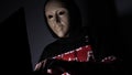 Male hacker hidden face with the mask accessing to personal information on laptop phone in the dark. Technology, cyber crime con