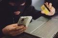 Male hacker in a robber mask uses phone, credit card and laptop in some fraudulent scheme. Cyber thief stole the personal data and Royalty Free Stock Photo