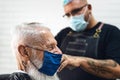 Male hairdresser cutting hair to hipster senior client while wearing face surgical mask - Young hairstylist working in barbershop