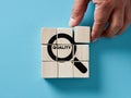 Male hand arranging wooden cubes with a magnifier focusing on the word quality. Quality control, searching for best quality