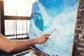 Male hand of artist holding paintbrush on background of canvas on easel. Professional painter drawing ocean wave in loft studio Royalty Free Stock Photo