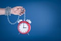 Male hand chained to red alarm clock on blue background