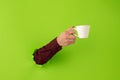 Male hand with cup of tea or coffee breaks through green paper background. Royalty Free Stock Photo