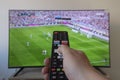 a male hand holding a black tv remote control with football game at background