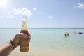 Male hand holding a bottle of beer against a sunny sky and crystal clear sea. In the background some people having fun in the Royalty Free Stock Photo