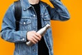 Male hand holding eco steel thermo bottle for water, wearing jeans jacket and backpack, on yellow background.