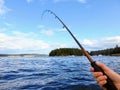 A male hand holding a fishing rod over the ocean. The rod is pointing right and upwards towards the sky. Room for text.