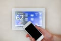 Male hand holding smart phone to operate heating or cooling via digital touch screen thermostat for home