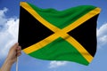 Male hand holds against the background of the sky with clouds the national flag of Jamaica on a luxurious texture of satin, silk