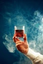 Male hand holds glass of whiskey, rum in smoke against gradient blue background with negative space to insert text. Royalty Free Stock Photo