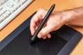 Male hand holds a pen on a black graphics tablet on a written wooden table