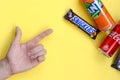 Male hand points on Snickers chocolate bar in brown wrapping, red Coca Cola and orange Fanta tin can on yellow pastel background. Royalty Free Stock Photo