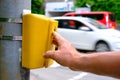 Male hand presses a yellow device with a button on demand with a symbol of a man to cross the road, a pedestrian observes the