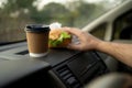 Male hand putting burger and coffee on panel, engaged in reckless eating and drinking while driving car