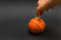 Male hand putting coin into the piggy bank with a shape of basket ball isolated on black background Royalty Free Stock Photo
