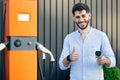 Male hand showing thumbs up holding power cable supply plugged at electric car charging station. Bearded man standing Royalty Free Stock Photo