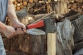 Male hands with an ax chop a wooden chock against the background of sawn logs and woodpile of firewood