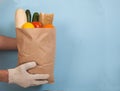 Male hands in cotton gloves hold a paper bag with pasta, tomatoes, cucumbers, daikon, lemon, orange on a blue background. Zero Royalty Free Stock Photo