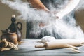 Male hands with flour clap over dough next to clay pot and oil bottle and rolling pin. Flour splash over dark table Royalty Free Stock Photo