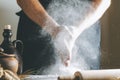 Male hands with flour clap over dough next to clay pot and oil bottle and rolling pin. Flour splash over dark table Royalty Free Stock Photo