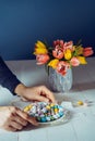 Male hands putting plate with Easter colorful sweet eggs on the white wooden table with fresh tulip flowers vase and dark blue Royalty Free Stock Photo