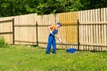 Male handyman wearing blue overalls paints wooden fence