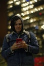 Male hipster with smartphone at night in city. Low angle of serious young male with dreadlocks browsing mobile phone