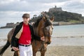 Handsome man, Male Horse Rider walking with his horse on beach, wearing traditional flat cap, white trousers, red polo shirt Royalty Free Stock Photo