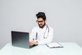 Male indian doctor wearing a white coat, sitting at a desk working with his laptop Royalty Free Stock Photo