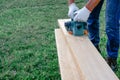 A male joiner planes a wooden plank on the lawn with an electric plane. DIY in the backyard. Handsome planing wooden plank with a