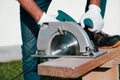 A male joiner works with a hand-held circular saw. A builder is sawing a board at the construction site of a new house