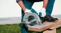 A male joiner works with a hand-held circular saw. A builder is sawing a board at the construction site of a new house