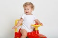 Male kid with wavy blond hair driving red and yellow toy car indoor, looking away, dresses white bodysuit, posing isolated over Royalty Free Stock Photo