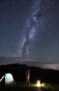 Male is looking at bonfire near tent under starry sky on which Milky Way is visible. Royalty Free Stock Photo