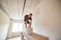 Male is making repairing in apartment by putting putty on the walls and standing on wooden stand against a window