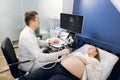 Male mature professional obstetrician, examining belly of pretty young pregnant woman by ultrasonic scan in modern Royalty Free Stock Photo