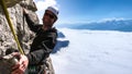 Male mountain guide smiling on a steep vertical rock climb in gorgeous surroundings high above a sea of clouds in the valley below