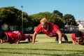 Male multiracial players in red uniforms practicing push-ups on grassy field against clear sky