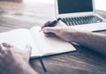 Male person writing in notebook besides laptop computer on wooden desk