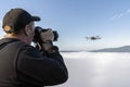 a male photographer takes pictures of a drone hovering in a thick fog in the air above the top of a mountain, while a