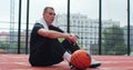 Male player sitting on basketball sports ground with ball. Concentrated sportsman looking at camera during training. Royalty Free Stock Photo