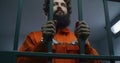 Male prisoner holds metal bars, stands in prison cell in handcuffs